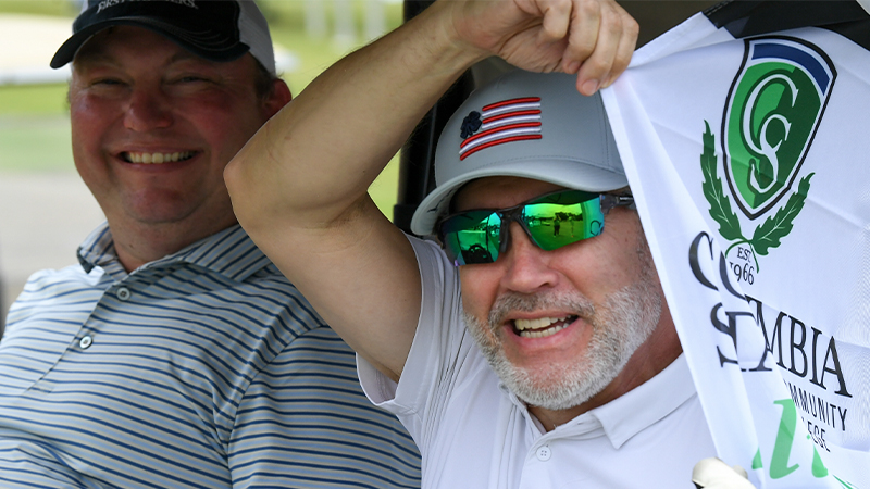 Two men sit in a golf cart, wearing a white shirt and sunglasses, holds up a flag with the logo of Columbia State